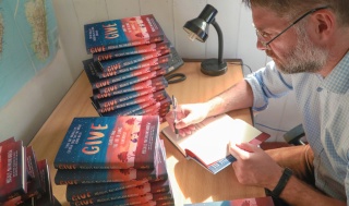 magnus macfarlane-barrow sitting at his desk signing a copy of his book, GIVE.