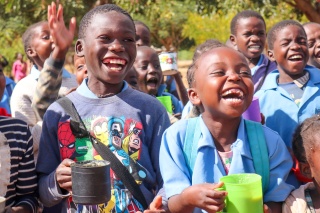 Image of children holding mugs filled with food and laughing, looking beyond the camera. 